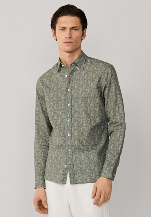 Homme portant une chemise verte à manches longues avec un motif abstrait blanc subtil, associée à un pantalon blanc, debout devant un fond uni.