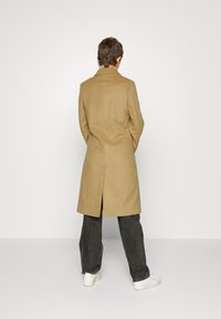 Cappotto lungo beige con un design strutturato e spacco sul retro, abbinato a jeans scuri e sneakers bianche. Estetica semplice e moderna.