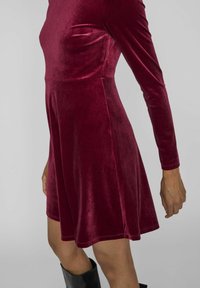 Kvinna som bär en långärmad, knälång, bordeauxvelourklänning med en figurnära midja och svarta knähöga stövlar mot en enfärgad bakgrund.
