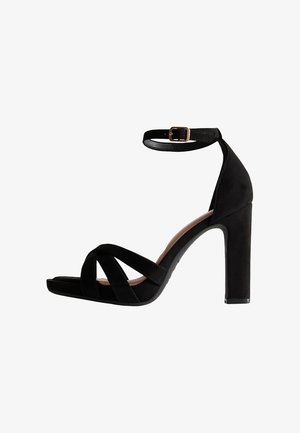 Sandalo alto in suede nero con cinturini incrociati, cinturino alla caviglia con fibbia dorata e tacco a blocco. La suola e la fodera sono di colore beige.