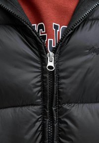 Doudoune noire avec une texture lisse, dotée d'une fermeture éclair à l'avant et d'accents côtelés noirs. Un sweat-shirt rouge est visible en dessous.