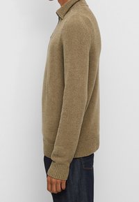 Pull en tricot beige avec un col, présentant une finition texturée, des poignets et un bas côtelés, et une coupe droite, porté sur un jeans noir.