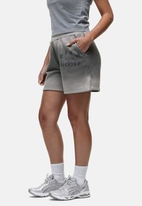 Pantaloni corti di cotone grigi con tasche laterali e testo stampato in grigio scuro. Indossati con una canotta grigia e scarpe da corsa argentate con calze bianche.