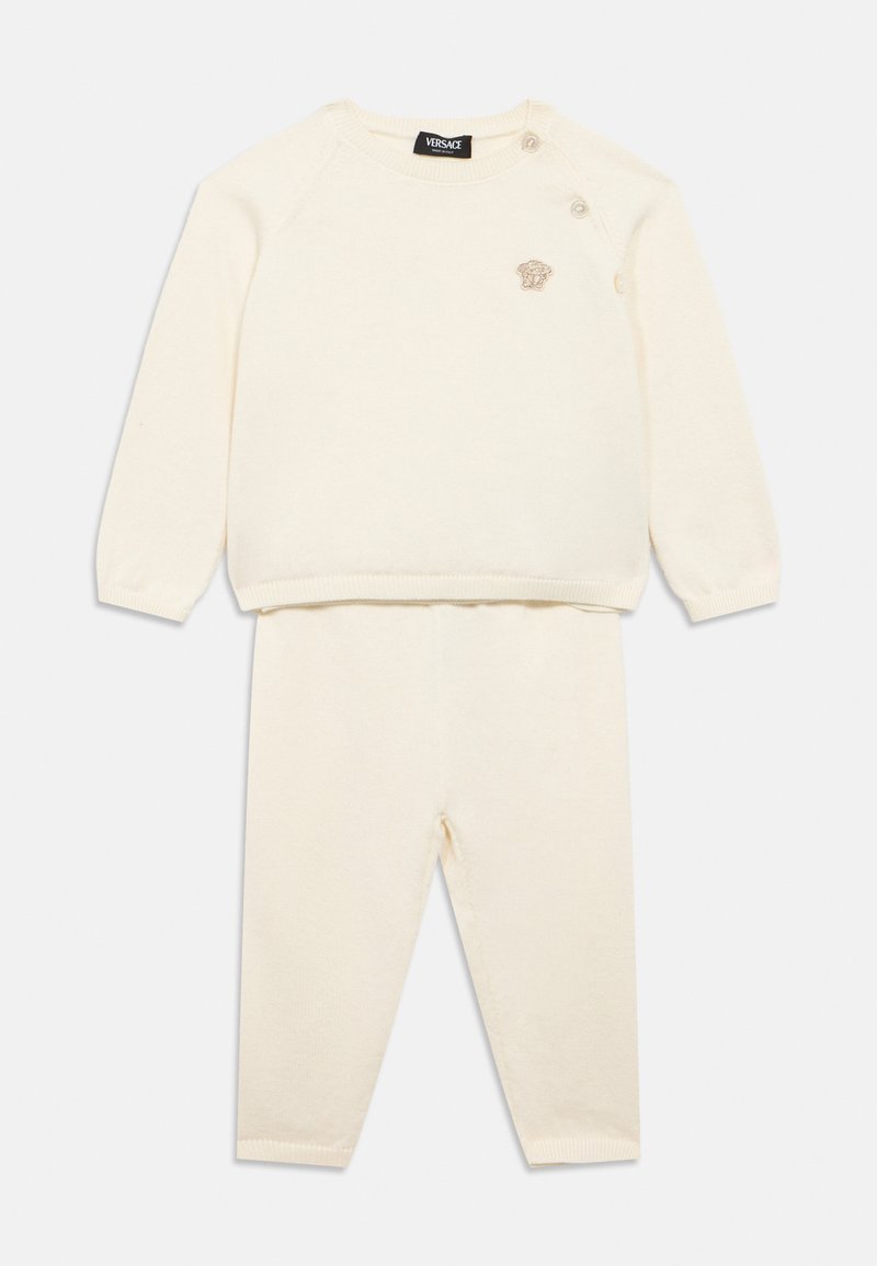 Σετ δύο τεμαχίων σε κρεμ χρώμα από πλεκτό ύφασμα, που περιλαμβάνει μπλούζα με μακριά μανίκια και κουμπιά στους ώμους και παντελόνι σε ίδιο ύφος, φτιαγμένο από απαλό ύφασμα.