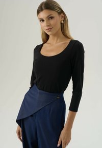 Jonge vrouw met lang blond haar, die gouden hangers draagt, een zwart shirt met lange mouwen en hoog gesloten marineblauwe broek met wikkel detail.