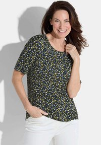 Korte mouwen marineblauw top met een gestippeld patroon in blauw, groen en wit, met een ronde halslijn en een relaxte pasvorm. Gecombineerd met witte broek.