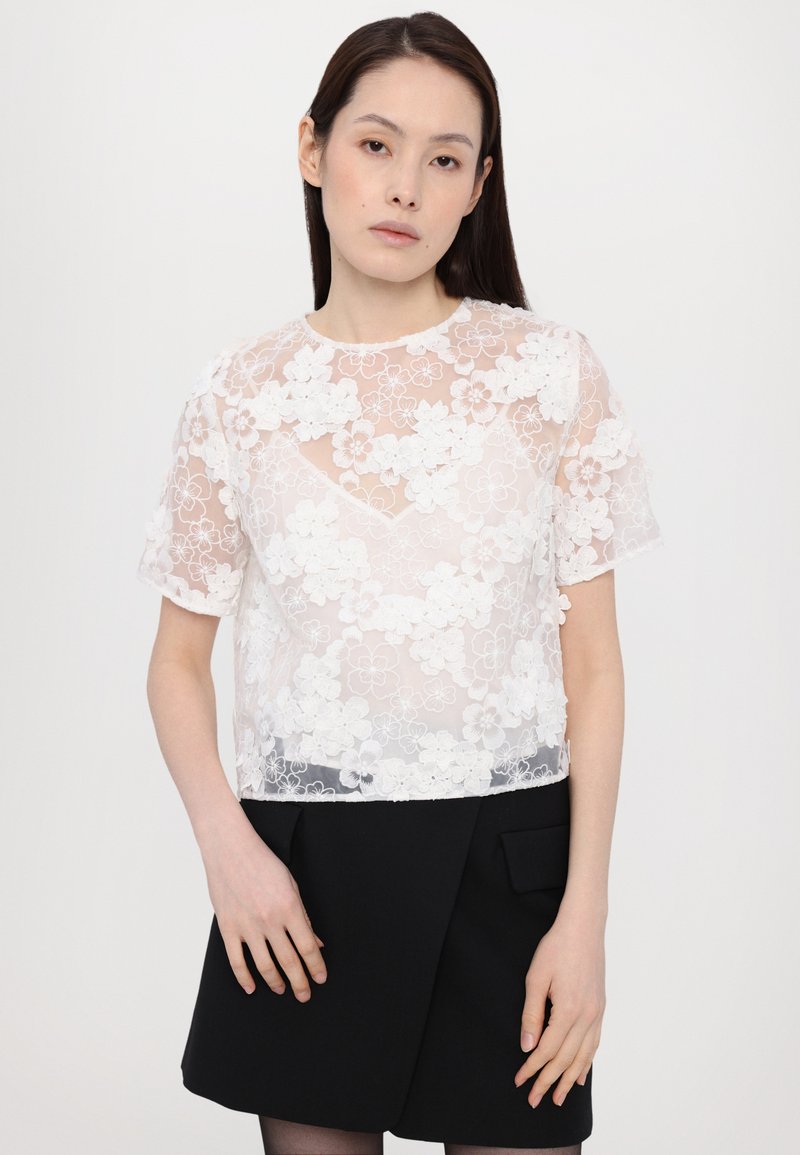 Witte bloemenkant top met korte mouwen en een transparant ontwerp. Heeft een ronde halslijn en gelaagde bloemdetails bovenop een effen zwarte rok.