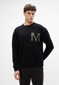 Suéter negro hecho de suave tejido de punto, con un gran emblema verde en forma de "M" en la parte delantera, y con cuello y puños acanalados.