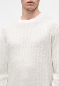 Jeune personne portant un pull en maille blanche texturée avec un motif losange subtil et un col rond bord-côtes.
