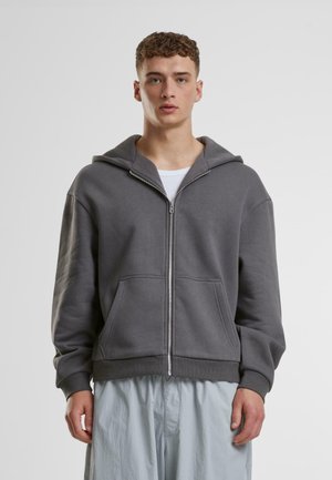 Grå zip-up hoodie lavet af blød stof, med en stor hætte, frontlommer og ribbede manchetter. Barbere over en hvid T-shirt.