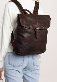 Bruine leren rugzak met een klepsluiting, textuur oppervlak, verstelbare banden en een metalen gesp accent. Draag op de rug tegen een lichte outfit.