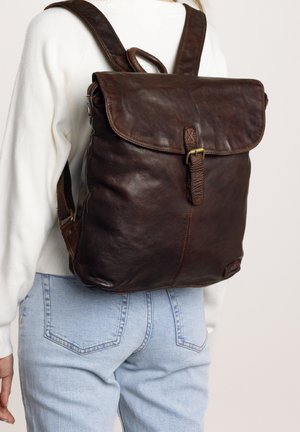 Brown leather backpack with a flap closure, textured surface, adjustable straps, and a metal buckle accent. Worn on the back against a light outfit.
