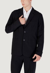 Uomo che indossa un giubbotto nero con due bottoni sopra una camicia bianca, mentre sistema il giubbotto vicino al petto.