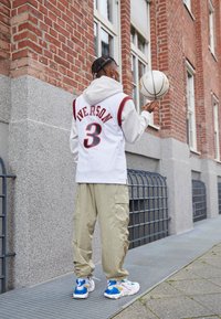 Henkilö, joka käyttää valkoista Allen Iversonin koripallopaitaa, vaaleita housuja ja tennareita, pyörittää valkoista koripalloa sormellaan tiiliseinän vieressä.