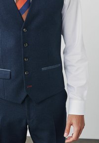 Marineblaue Weste mit einem strukturierten Muster, ausgestattet mit fünf braunen Knöpfen, zwei Taschen und einem subtilen roten Akzent am Saum. Ein weißer Ärmel ist sichtbar.