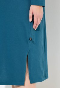 Vestido de algodón en color verde azulado con mangas largas, aberturas laterales y una pequeña etiqueta de tela en el dobladillo. Textura suave y diseño sencillo.