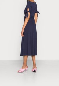 Robe bleu marine longueur genou avec manches courtes ornées de liens. Associée à des talons ouverts roses à talon bloc.