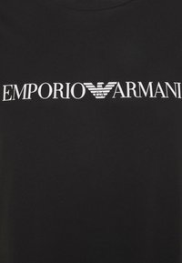 Mustassa puuvillapaidassa on selkeä valkoinen "Emporio Armani" -logo, jonka yläpuolella on tyylitelty kotka-embleemi. Pehmeä pinta.