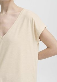 Prikaz osobe u bež majici s kratkim rukavima i V-izrezom, s desnom rukom savijenom iza leđa, na jednobojnoj pozadini.