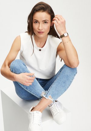Witte mouwloze top, lichtblauwe ripped jeans en witte sneakers. Accessories omvatten een dunne armband en een horloge met een groene band.