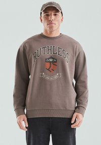 Młody mężczyzna z piegami, noszący brązową czapkę i brązowy sweatshirt z grafiką "RUTHLESS" i tarczą, stoi na tle jasnym.