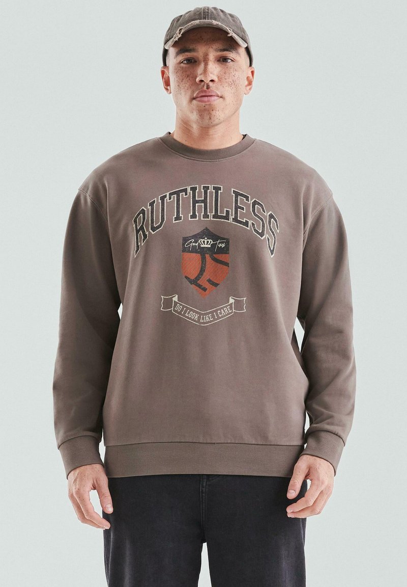 Młody mężczyzna z piegami, noszący brązową czapkę i brązowy sweatshirt z grafiką "RUTHLESS" i tarczą, stoi na tle jasnym.