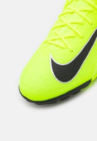 Botín de fútbol de un brillante color amarillo neón con tela texturizada, logo de Nike en negro, y un diseño de cordones intrincado. Suela negra con un patrón de tracción.