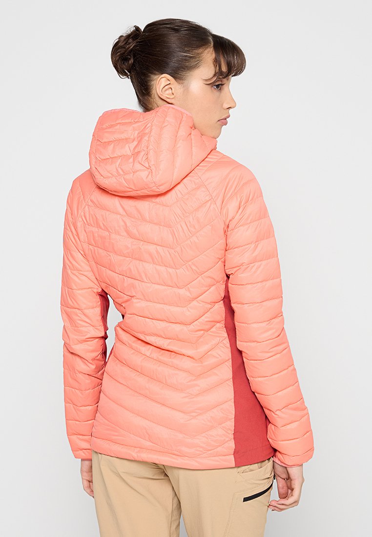 Mujer con cabello castaño recogido, vestida con una chaqueta acolchada color coral con capucha, mirando de lado, pantalones beige con bolsillo lateral, contra un fondo blanco.