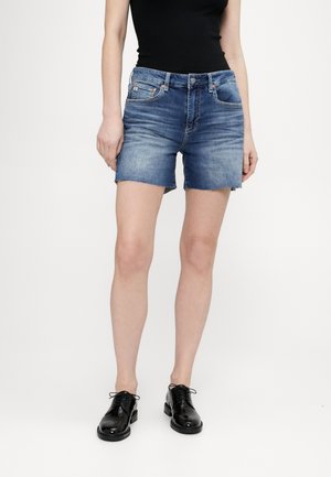 Shorts in denim blu medio, con un classico design a cinque tasche, orlo sfilacciato e silhouette aderente, abbinati a scarpe in pelle nera.