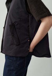 Eine Person trägt eine schwarze gepolsterte Weste mit einer Druckknopf-Tasche, einen dunklen, kurzärmeligen Pullover und blaue Jeans, mit der Hand in der Jeans-Tasche.
