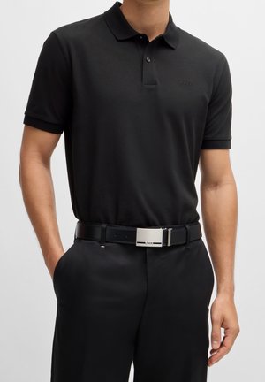 Homme portant un polo noir à manches courtes rentré dans un pantalon noir avec une ceinture en cuir noire équipée d'une boucle argentée.