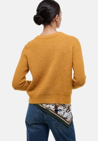 Pull en maille jaune moutarde avec une finition texturée. Le dos présente un ourlet en écharpe à motifs avec des accents blancs, marron et verts, associé à un jean bleu.