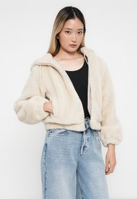 Krém színű műszőrme cropped dzseki kapucnival, húzózsinórral és elülső cipzárral. Fekete toppal és világoskék, bő szárú farmernadrággal párosítva.