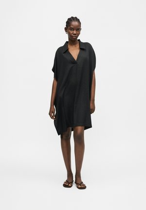 Sieviete ar pītu matu sakārtojumu, valkājot vaļīgu melnu V veida kakla kleitu un brūnas sandales, stāvot vienkārša balta fona priekšā.