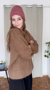 Barna bolyhos pulóver laza szabással, rózsaszín kötött sapkával párosítva. A háttérben bézs függönyök és egy zöld növény látható.