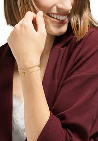 Bracciale doppio strato dorato con dettagli a forma di cuore, caratterizzato da catene delicate. Indossato su un polso accostato a una giacca borgogna profondo.