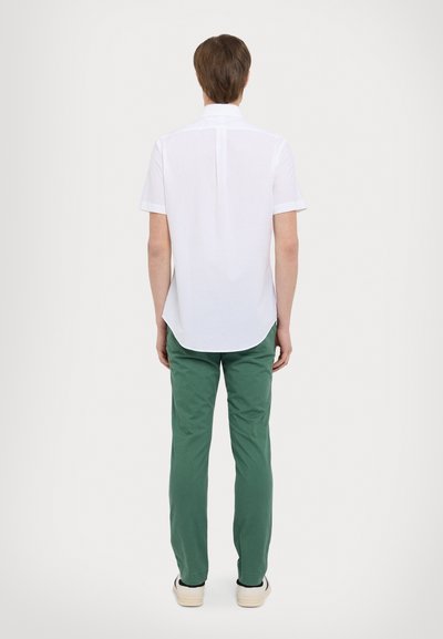 Chemise blanche à manches courtes, de coupe droite, associée à un pantalon vert, vue de dos. Présente un col plat et un ourlet arrondi.