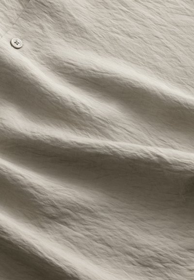 Ljusbeige tyg med en texturerad, något skrynklig yta och subtila mönster. En enkel vit knapp syns nära kanten.