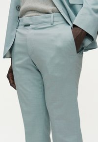 Άνδρας που φοράει ανοιχτό μπλε παντελόνι κοστουμιού και μπλε σακάκι, με το χέρι στην τσέπη, σε λευκό φόντο.