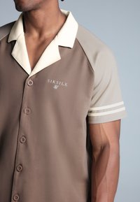 Smeđa majica kratkih rukava s kragnom u boji kreme, koja ima SIKSILK logo i prugaste detalje na rukavima. Glatka tekstura tkanine.