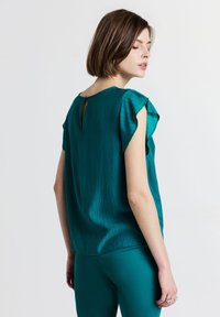 Donna che indossa una blusa corta a maniche corte di colore teal con scollo a chiave e pantaloni abbinati dello stesso colore, che guarda sopra la spalla su uno sfondo semplice.