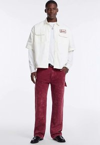 Weißes Hemd mit kurzen Ärmeln und Fronttaschen, kombiniert mit rot eingefärbter Jeans mit dekorativen Nähten und einem lockeren Sitz. Schwarze Schuhe runden den Look ab.