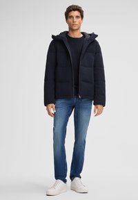 Doudoune marine avec capuche, dotée d'un design matelassé. Portée sur un pull en maille marine, associée à un jean bleu et des baskets blanches.