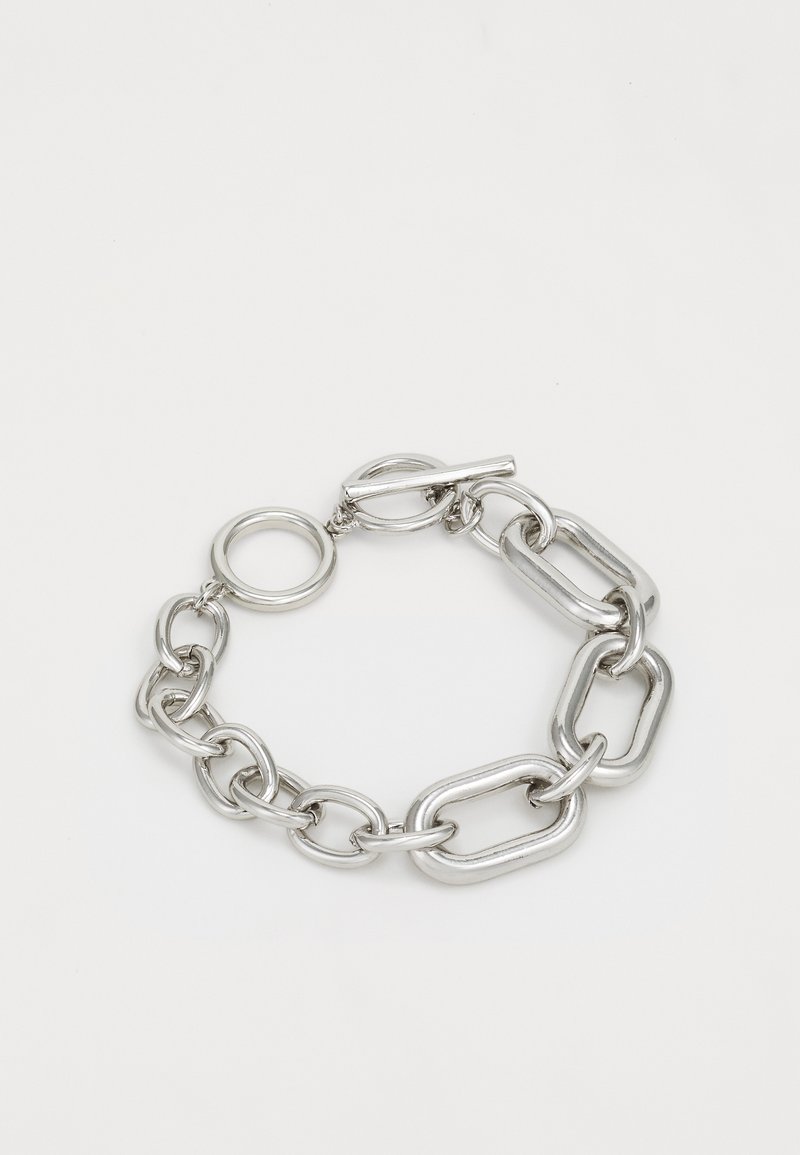 Pulseira de corrente em prata com elos ovais e retangulares entrelaçados, equipada com um fecho de palhetas e um anexo de medalha circular. Textura suave.