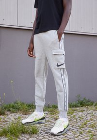 Ljusgrå sweatpants med sidoficka för cargo, svart Nike-logotyp, svarta sidostygn och ribbade muddar. Matchas med vita och gröna sneakers.