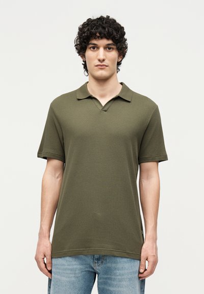 Jeune homme aux cheveux bouclés et foncés portant un polo à manches courtes vert olive et un jean bleu clair sur un fond uni.
