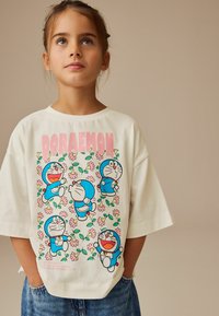 Fehér pamut póló, színes "Doraemon" nyomattal, különböző pózokban és virágos díszítéssel. Lozsasan szabott, rövid ujjú. Kék és rózsaszín részletekkel.