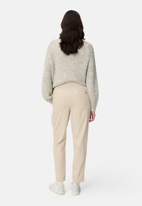 Femme aux longs cheveux foncés portant un pull en maille beige, un pantalon beige clair avec des poches arrière, et des baskets blanches, debout de dos.