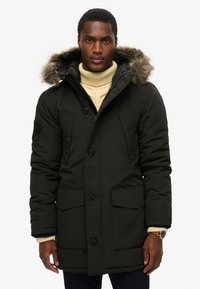 Zwarte gewatteerde parka-jack met een met bont gevoerde capuchon, twee voorkruiszakken, knoopsluiting en geribbelde manchetten; gedragen over een crèmekleurige coltrui.