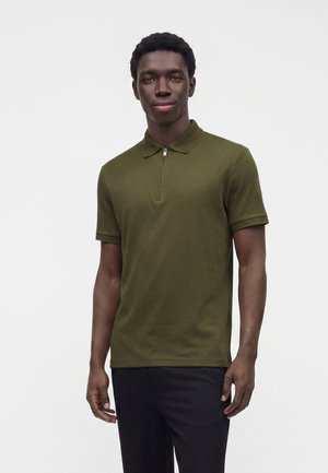 Homme debout devant un fond uni, portant un polo vert olive à manches courtes avec un col zippé et un pantalon noir, tourné vers l'avant.